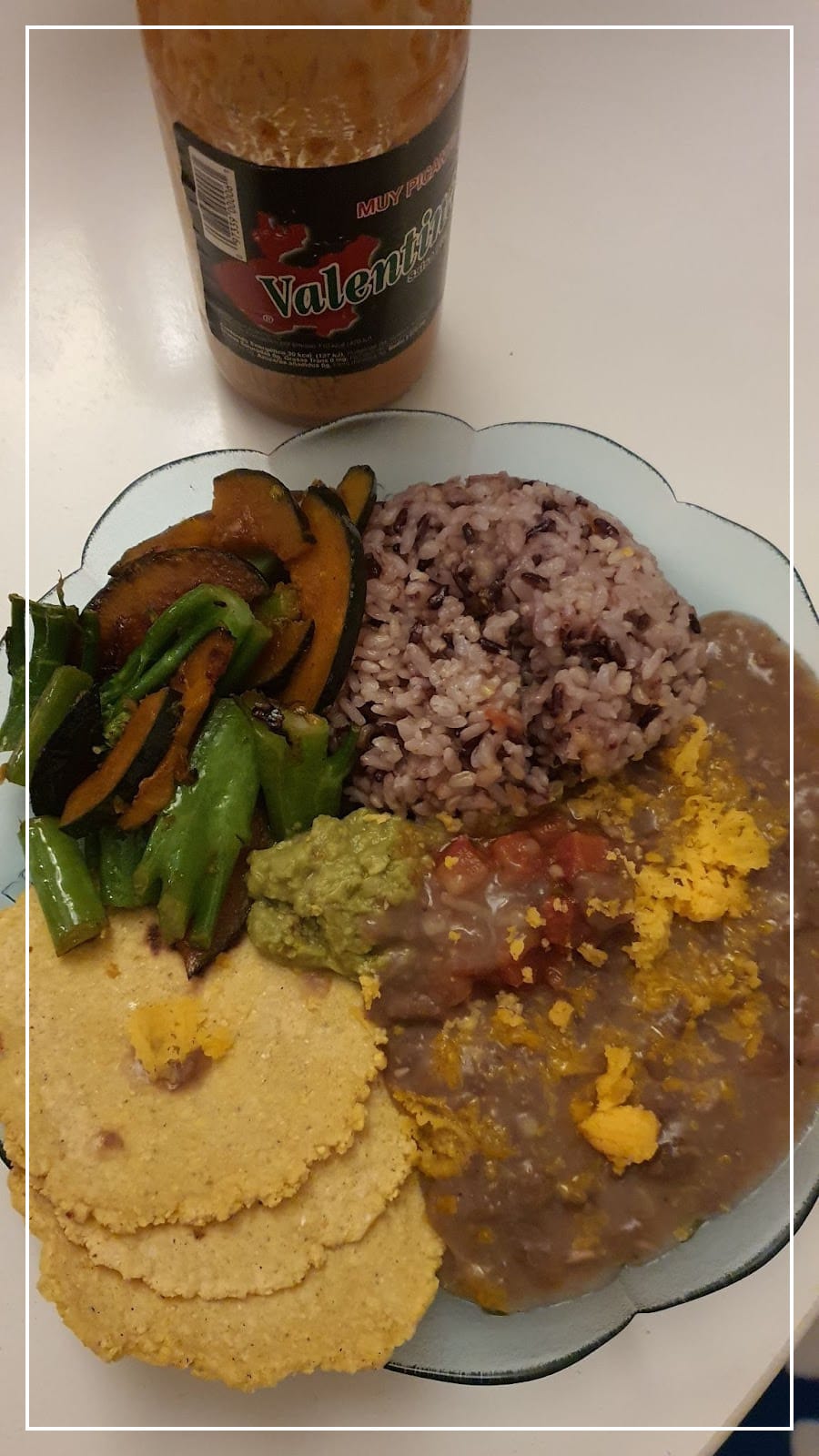 a plate of refried beans served with handmade corn tortillas, salsa, guacamole, leftover vegetables, and mixed rice. Next to the plate is a bottle of Valentina hot sauce.